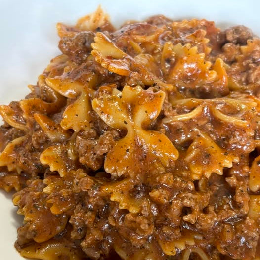A close-up shot of a serving of Epicure Skillet Lasagna made with bowtie pasta in a rich meat and tomato sauce, served on a white plate.