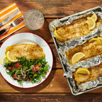 Three cooked fish fillets on aluminum foil with lemon slices, served on a wooden table with a side salad and another plated fillet with salad.