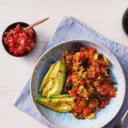 A styled breakfast scene on a light wood table featuring a plate of colorful Spanish Rice Migas, scrambled eggs with avocado, tomatoes, and peppers, a cup of black coffee, a bowl of salsa, and toasted bread on a blue napkin.