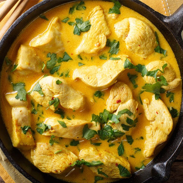 A cast iron skillet filled with a Stovetop Coconut Chicken  garnished with chopped green herbs, set on a rustic wooden table with wooden spoons and a napkin.