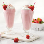 Two pink strawberry milkshakes in tall clear glasses, made with Epicure Strawberry Margarita Mix, topped with whipped cream, a strawberry garnish, and pink sprinkles, arranged on a marble board with a small bowl of sprinkles and a basket of strawberries on a light marble surface.