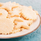Star-shaped and round sugar cookies dusted with powdered sugar, piled on a white ceramic plate with a gold rim against a textured light blue background.