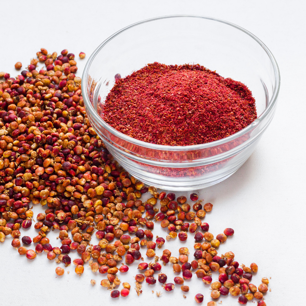 A glass bowl filled with ground sumac and a pile of sumac berries spilled on the side.