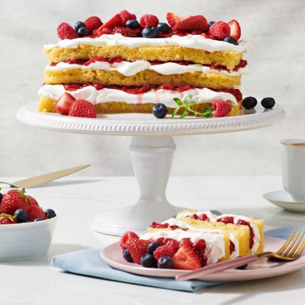 Layered berry cake on a white cake stand with a slice on a plate, accompanied by a cup of tea.
