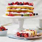 Layered berry cake on a white cake stand with a slice on a plate, accompanied by a cup of tea.
