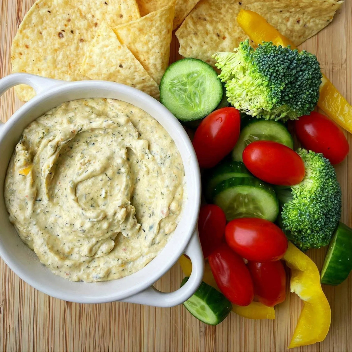 A white bowl filled with creamy herb dip sits on a wooden board, surrounded by tortilla chips, broccoli florets, sliced cucumbers, cherry tomatoes, and yellow bell pepper strips.