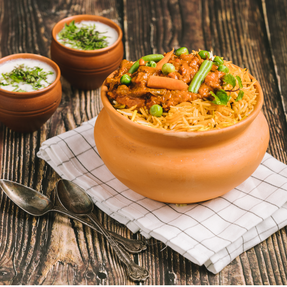 A terracotta clay pot filled with vegetable biryani, set on a white checkered cloth over a rustic wooden table. Two small bowls of yogurt with chopped herbs are blurred in the background.