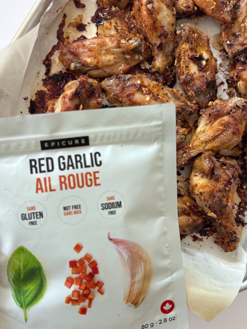 A white pouch of Epicure Red Garlic Topper placed in front of cooked chicken wings on parchment paper. The package shows the brand Epicure and product name Red Garlic Ail Rouge, with illustrations of a basil leaf, diced red pepper, garlic, and a red maple leaf.
