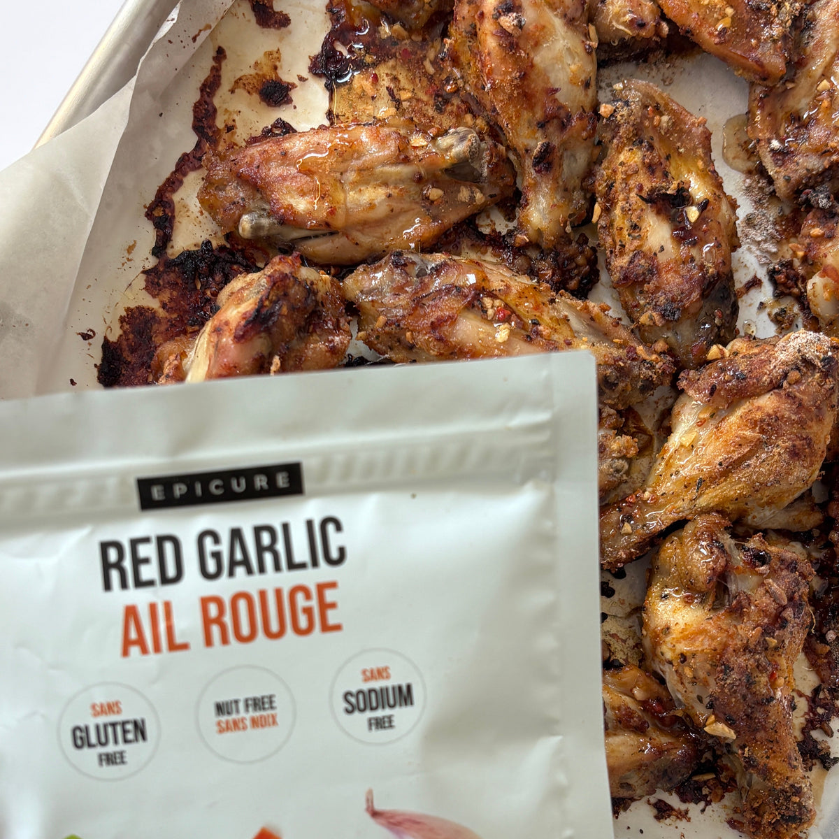A white pouch of Epicure Red Garlic Topper placed in front of cooked chicken wings on parchment paper. The package shows the brand Epicure and product name Red Garlic Ail Rouge, with illustrations of a basil leaf, diced red pepper, garlic, and a red maple leaf.