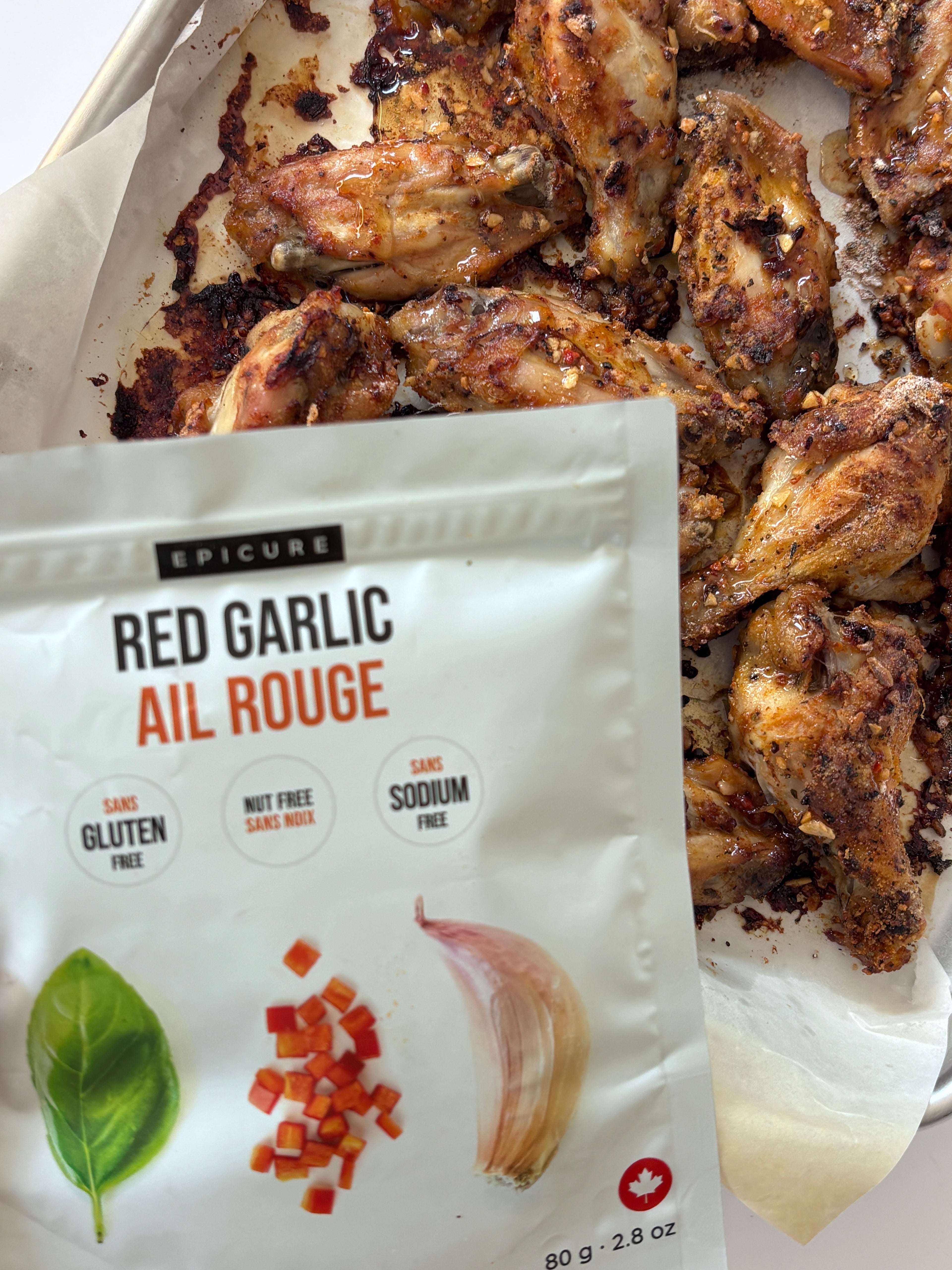 A white pouch of Epicure Red Garlic Topper placed in front of cooked chicken wings on parchment paper. The package shows the brand Epicure and product name Red Garlic Ail Rouge, with illustrations of a basil leaf, diced red pepper, garlic, and a red maple leaf.