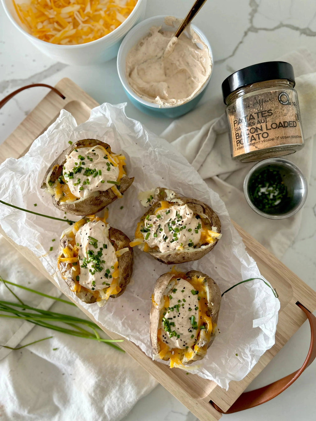 Bacon Loaded Potato Vegan Seasoning jar beside four loaded baked potatoes topped with vegan sour cream shredded cheese and chopped chives on a wooden board