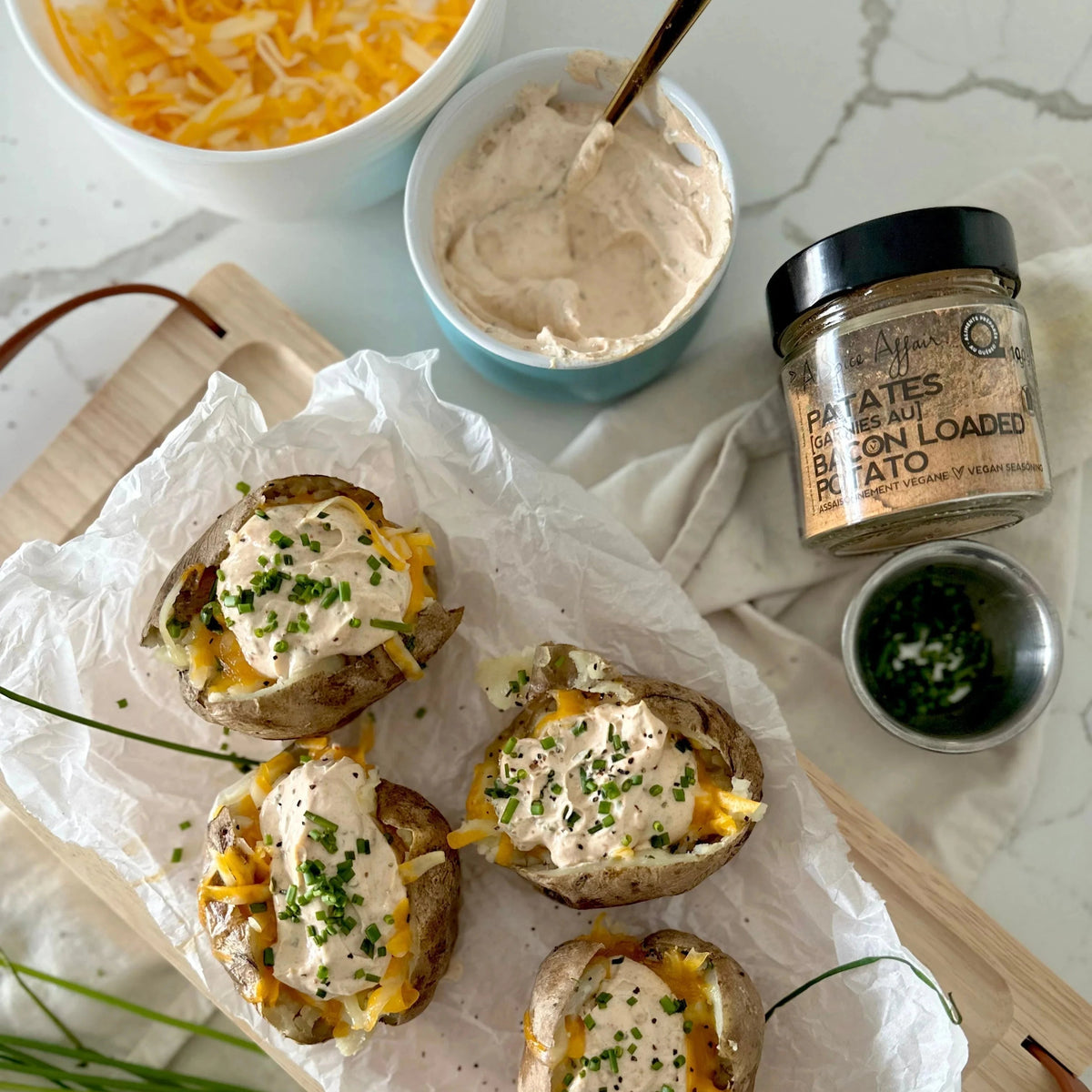 Bacon Loaded Potato Vegan Seasoning jar beside four loaded baked potatoes topped with vegan sour cream shredded cheese and chopped chives on a wooden board