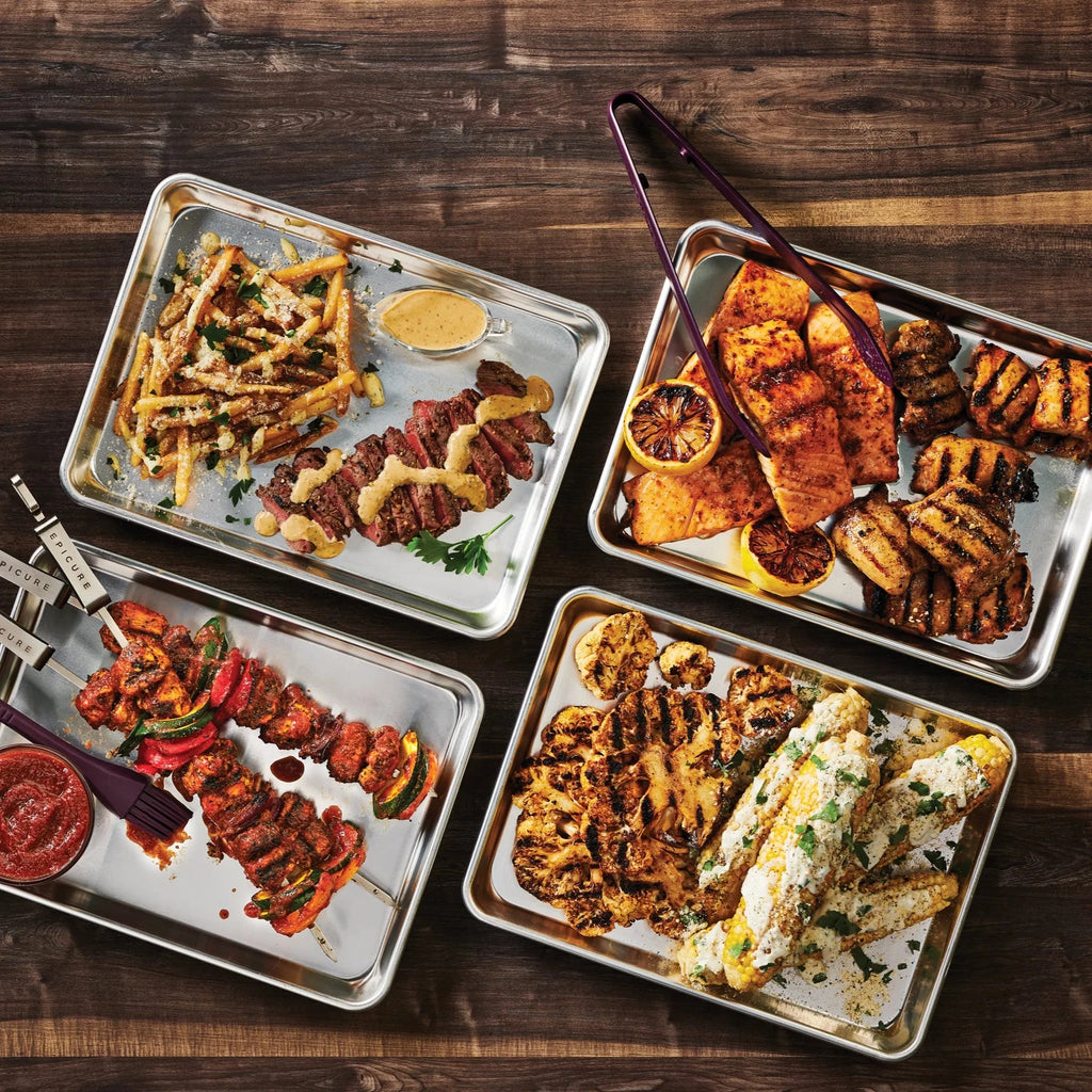 Four trays of grilled and roasted meats and vegetables on a wooden surface, seasoned with Epicure BBQ Chicken & Rib Rub