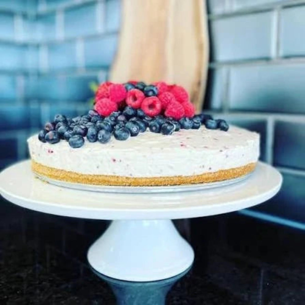 Epicure Berry Swirl No-Bake Cheesecake Mix prepared as a creamy berry cheesecake with a golden crumb crust topped with fresh blueberries and raspberries on a white cake stand