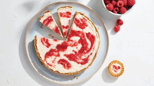 Berry Swirl No-Bake Cheesecake Mix featuring a creamy raspberry-swirled cheesecake on a plate, sliced into wedges with fresh raspberries and a mini tart nearby