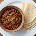 A bowl of chili with beans, meat, diced tomatoes, and cilantro garnish, served on a white plate with folded tortillas.