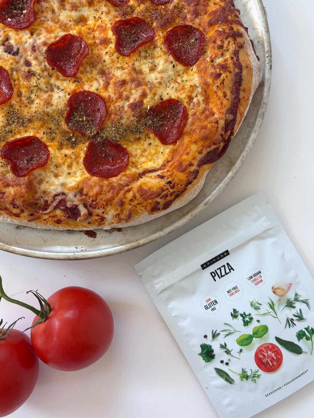 A close-up of a pepperoni pizza on a metal pan, styled with two red tomatoes and a white pouch of Epicure pizza seasoning on a white surface.