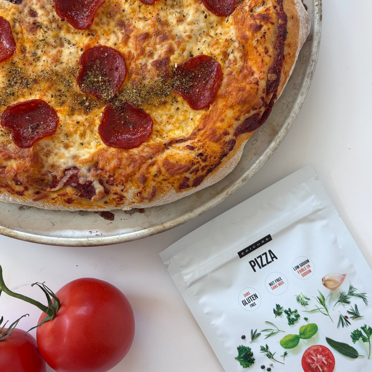 A close-up of a pepperoni pizza on a metal pan, styled with two red tomatoes and a white pouch of Epicure pizza seasoning on a white surface.