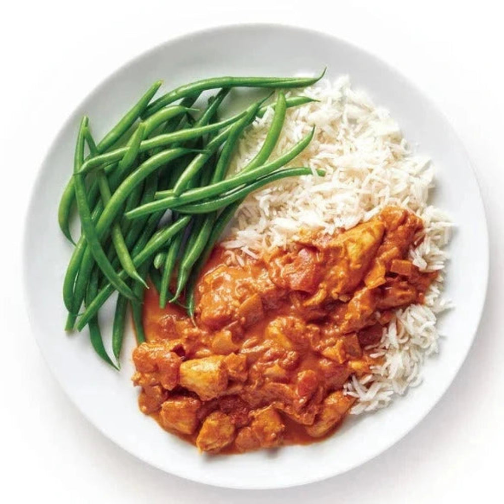  A white plate containing a serving of butter chicken on white rice with green beans on the side sitting on a white background.