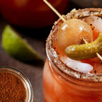 Caesar Rimmer & Seasoning beside a tomato cocktail with a seasoned glass rim, lime wedge, pickled garnish, skewer, and small bowl of spice blend