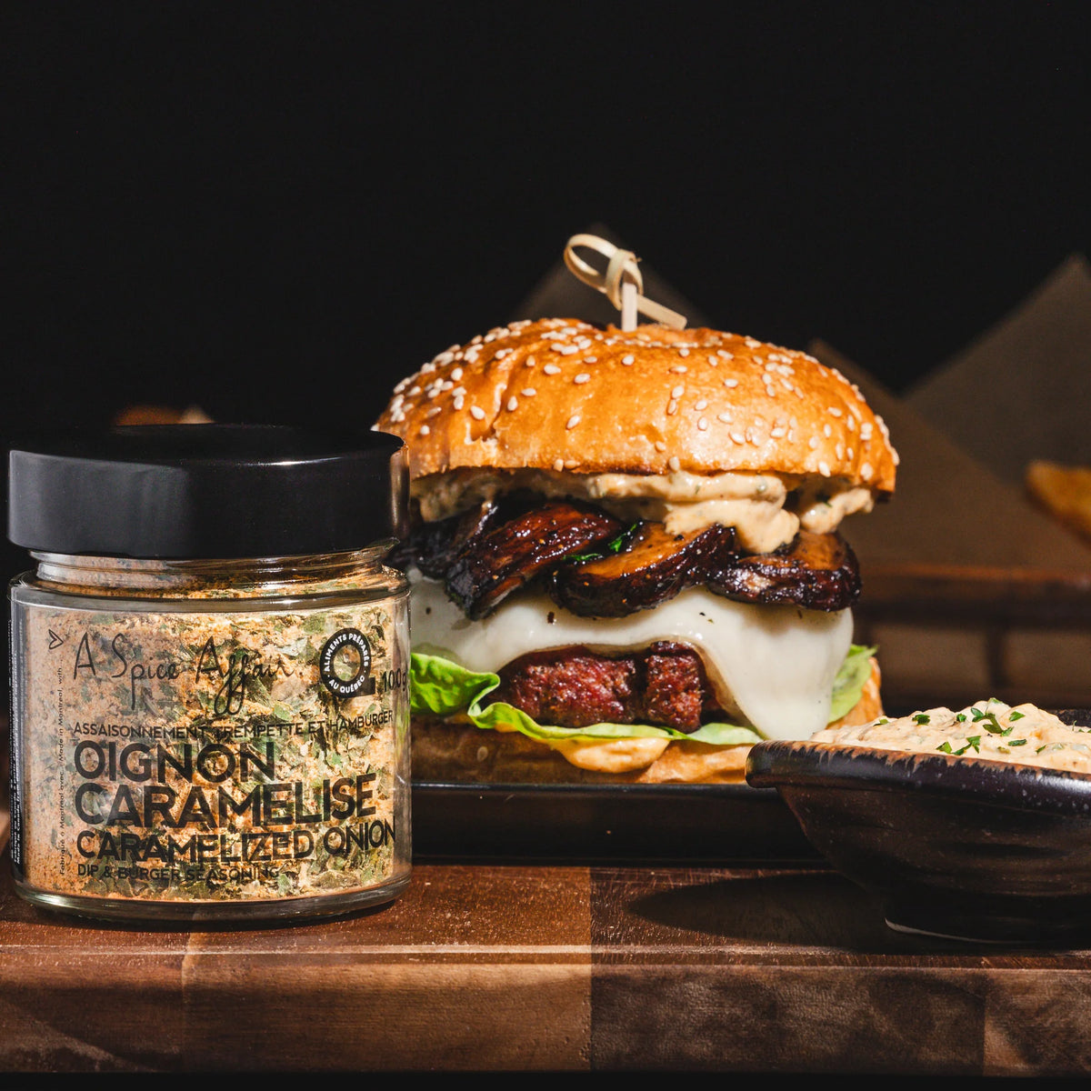 A jar of caramelized onion seasoning sits on a wooden board next to a gourmet burger with caramelized onions, melted cheese, and lettuce, with a side of coleslaw on a dark background.