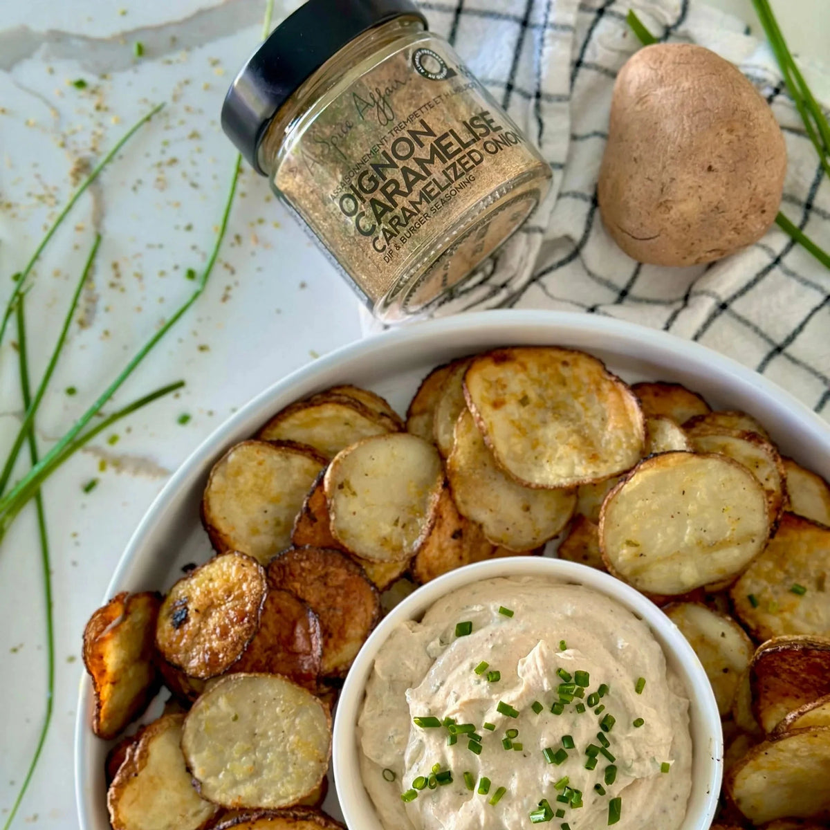 Bowl of roasted potato chips with a container of caramelized onion dip on a checkered cloth.