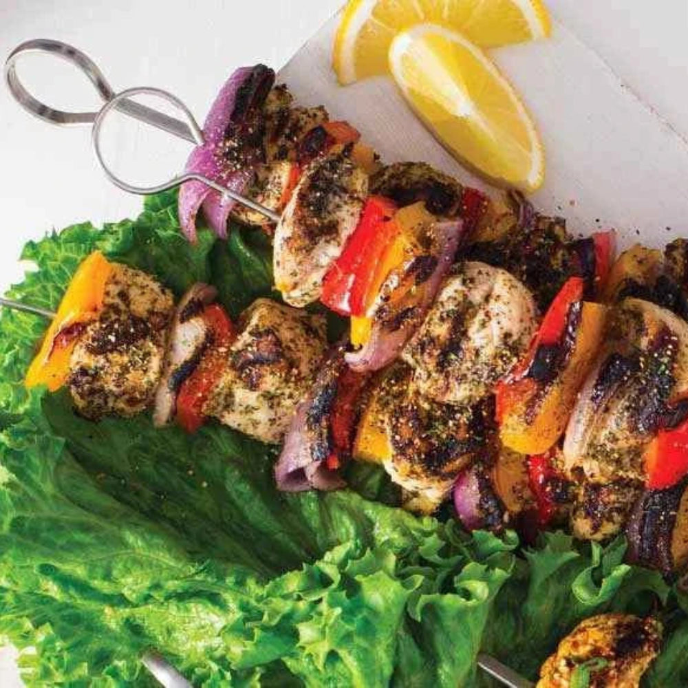 Skewer of grilled vegetables and meats on a bed of lettuce with lemon wedges and a purple drink.