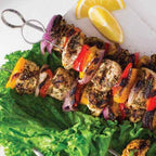 Skewer of grilled vegetables and meats on a bed of lettuce with lemon wedges and a purple drink.