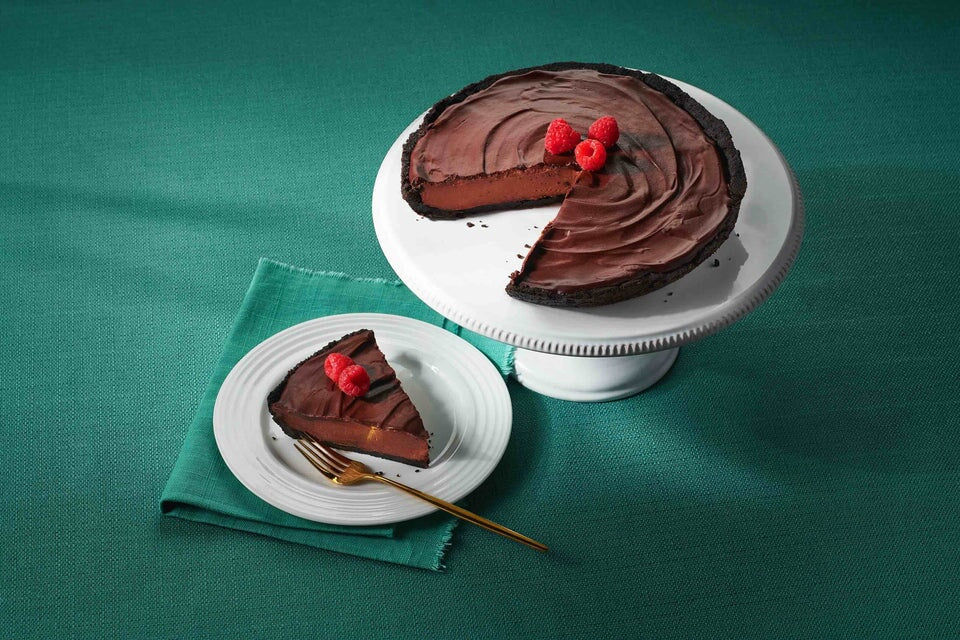Chocolate Velvet Cheesecake topped with raspberries on a white cake stand alongside a plated slice with fork on a teal cloth