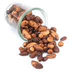 Crispy roasted almonds and cashews spilling out of a clear glass jar on a solid white background.