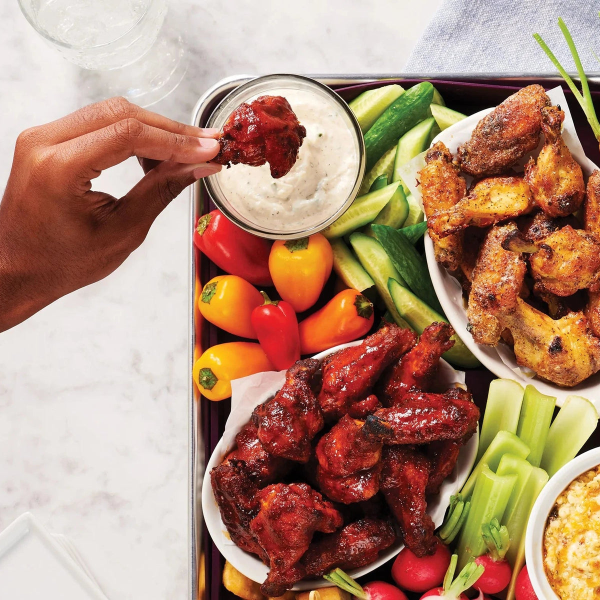 A tray of chicken wings, sliced vegetables and two bowls of dip on a white countertop 