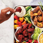 A tray of chicken wings, sliced vegetables and two bowls of dip on a white countertop 