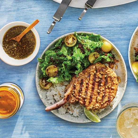 Two plated grilled pork chops with green salad, halved green cherry tomatoes, and lime wedges on a light blue wooden table