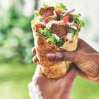 Hand holding a wrap with vegetables and donair meat against a blurred green outdoor background