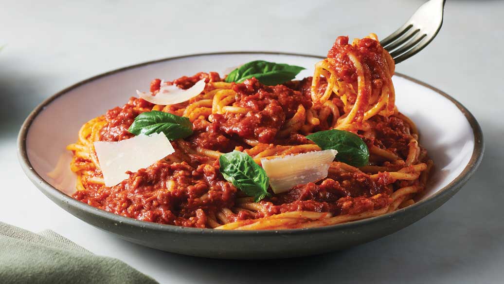 A close-up of a bowl of spaghetti with a rich red plant-based sauce, topped with fresh green basil leaves and shavings of parmesan cheese, with a fork lifting a portion of the pasta.