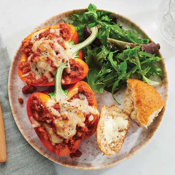Overhead view of a plated meal on a light surface: two halves of a red bell pepper stuffed with plant-based sauce and melted cheese, served with a side of green salad, a piece of bread on a white background
