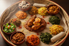 A large woven tray filled with an Ethiopian-style meal on a white injera cloth, featuring a central chicken and potato dish in red sauce, surrounded by side salads, yellow and green vegetables, and rolled flatbreads.