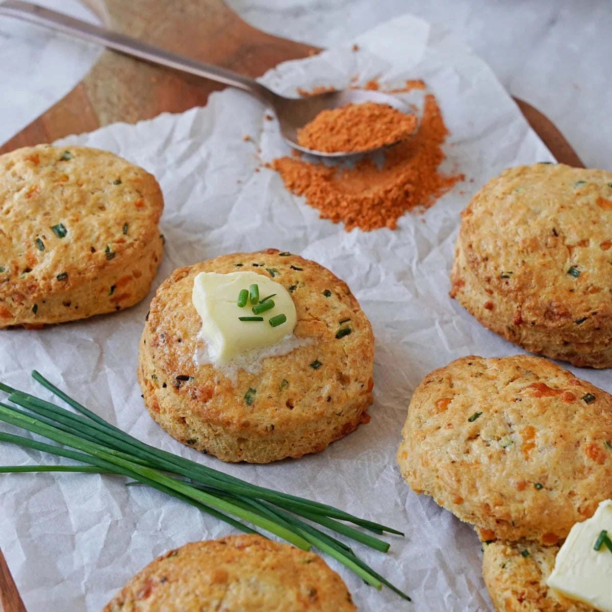 Everything Bacon Vegan Seasoning beside golden savory biscuits topped with chives and vegan butter on parchment with fresh green onions and a spoonful of seasoning