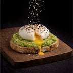 Avocado toast with poached egg on top being sprinkled with Everything Bagel seasoning sitting on a cutting board