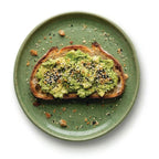 Everything Bagel Whole Food Topper sprinkled over mashed avocado toast on a rustic green plate with sesame seeds and herbs scattered around it