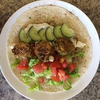 A top-down view of a plate of falafels on a tortilla wrap, garnished with cucumber slices, tomatoes, lettuce, and feta cheese, set on a speckled granite countertop.