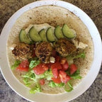 A top-down view of a plate of falafels on a tortilla wrap, garnished with cucumber slices, tomatoes, lettuce, and feta cheese, set on a speckled granite countertop.