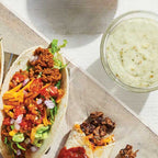 Three loaded soft tacos filled with ground meat, shredded cheese, diced tomatoes, onions, and lettuce, served on a wooden board next to a small glass bowl of Epicure Feisty Green Sauce on a white tabletop.