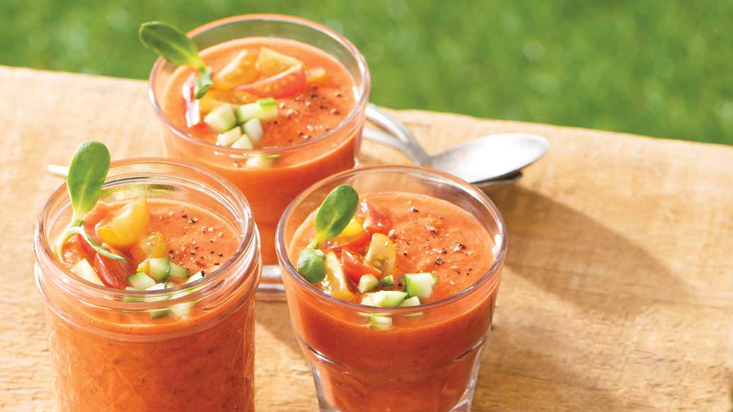 Three clear glass cups filled with red-orange gazpacho, garnished with diced vegetables and a small green leaf, arranged on a wooden board outdoors with a spoon in the background.