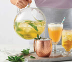 Person pouring Ginger Mule Herbal Tea Blend by Epicure from a glass pitcher into glasses on a wooden tray.