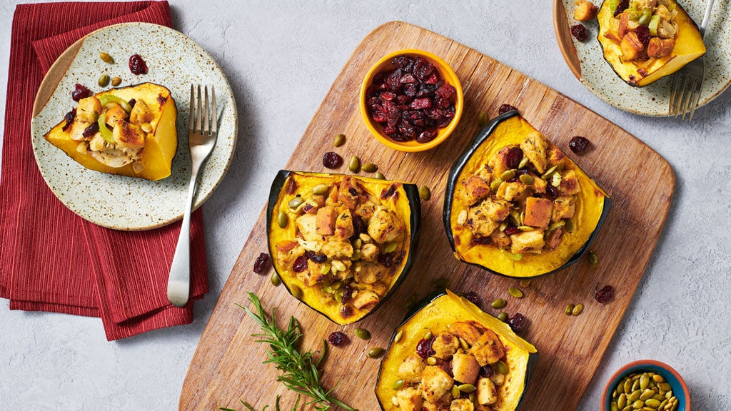 Stuffed acorn squash halves filled with prepared Epicure Happy Holiday Stuffing Mix, served on a wooden cutting board with garnish, alongside plates and a small bowl of cranberry sauce on a light gray surface.