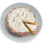 A round birthday cake with white frosting, colorful sprinkles around the edge, and five lit candles, shown on a light gray plate against a white background with one slice cut out.