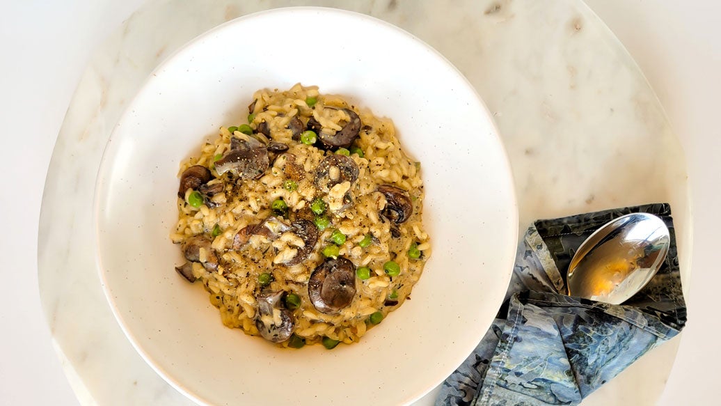 A top-down view of a creamy  Epicure Herb & Mushroom risotto in a white bowl, placed on a white marble plate with a folded blue napkin and a spoon.
