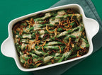 A white rectangular baking dish filled with green beans topped with melted cheese and crispy fried onions, set on a dark green surface with a folded gray napkin.