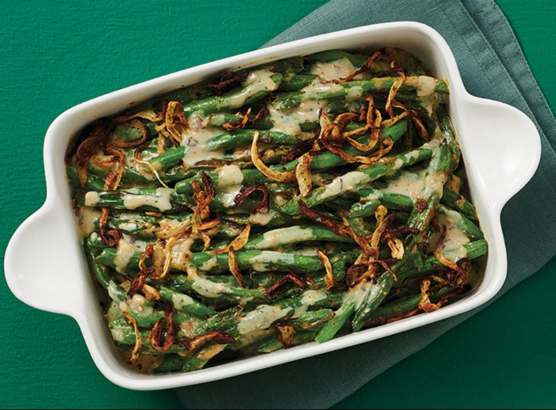 A white rectangular baking dish filled with green beans topped with melted cheese and crispy fried onions, set on a dark green surface with a folded gray napkin.